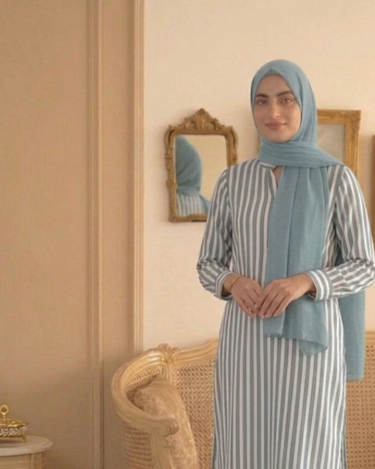 Woman in a striped outfit with a hijab standing in a room with a couch and decorative items.