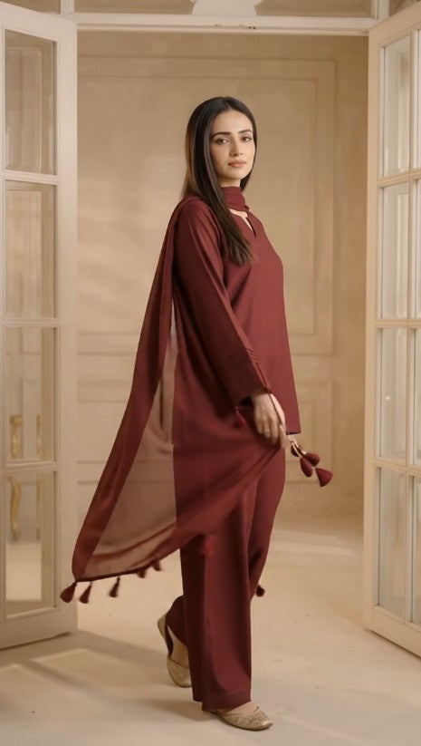 A woman ware ghazi fabric s  three-piece maroon traditional outfit with a matching sheer dupatta, standing in a warm, beige-toned room with open doorways.