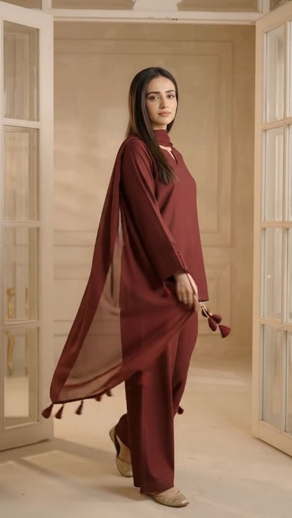 A woman ware ghazi fabric s  three-piece maroon traditional outfit with a matching sheer dupatta, standing in a warm, beige-toned room with open doorways.